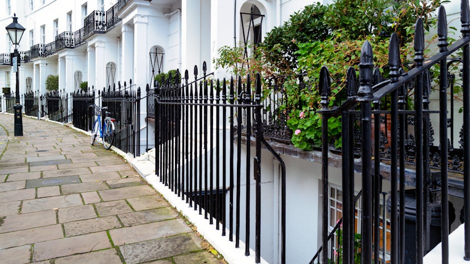 A narrow cobblestone street in a residential conservation area, flanked by white and beige terraced houses with black iron balconies and window trim. The street is lined with potted plants and small t